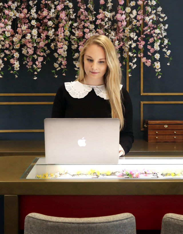 A salesperson in the stephen einhorn showroom looking at a laptop at the jewellery counter. There is a floral display behind her.